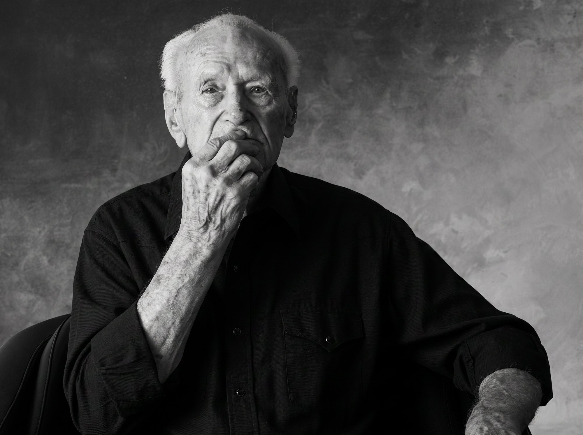 Editorial B&W portrait of an elderly man in a black western shirt, hand to chin, grey seamless backdrop, by G. John Slagle.