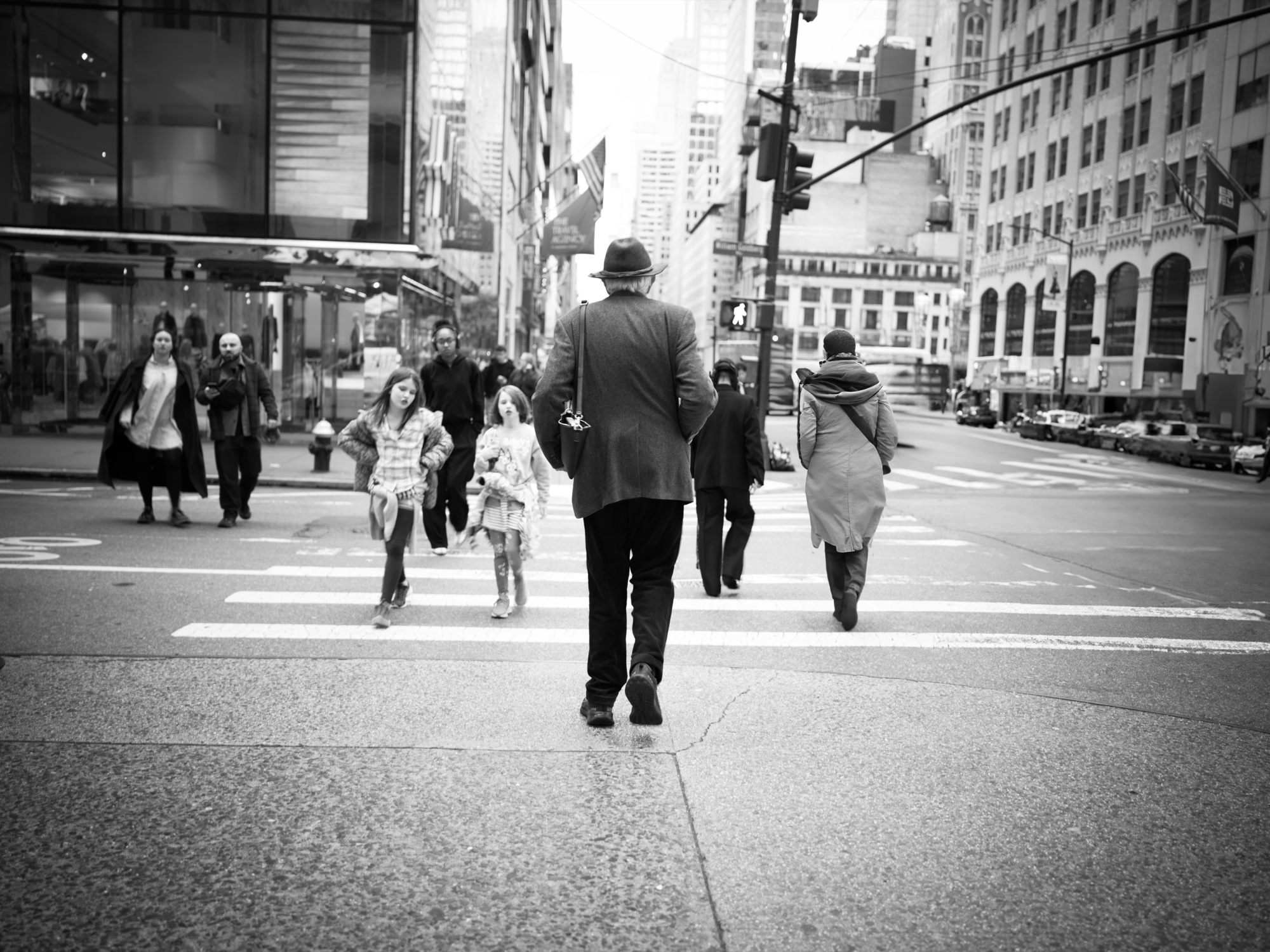 Man in fedora crosses a Manhattan avenue with two girls trailing behind, B&W.