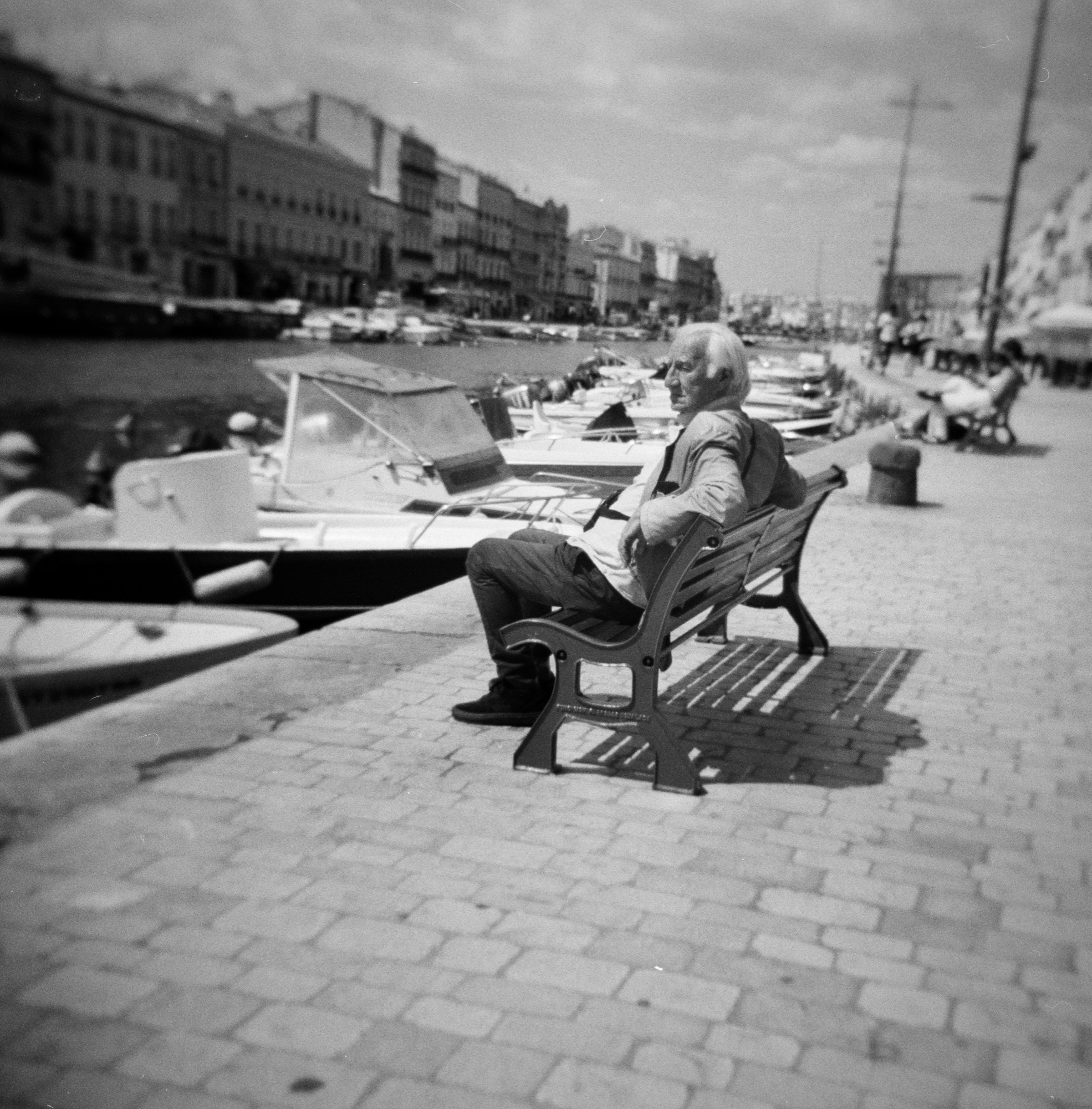 Older man on a bench beside boats moored along the harbor, S&egrave;te, France.