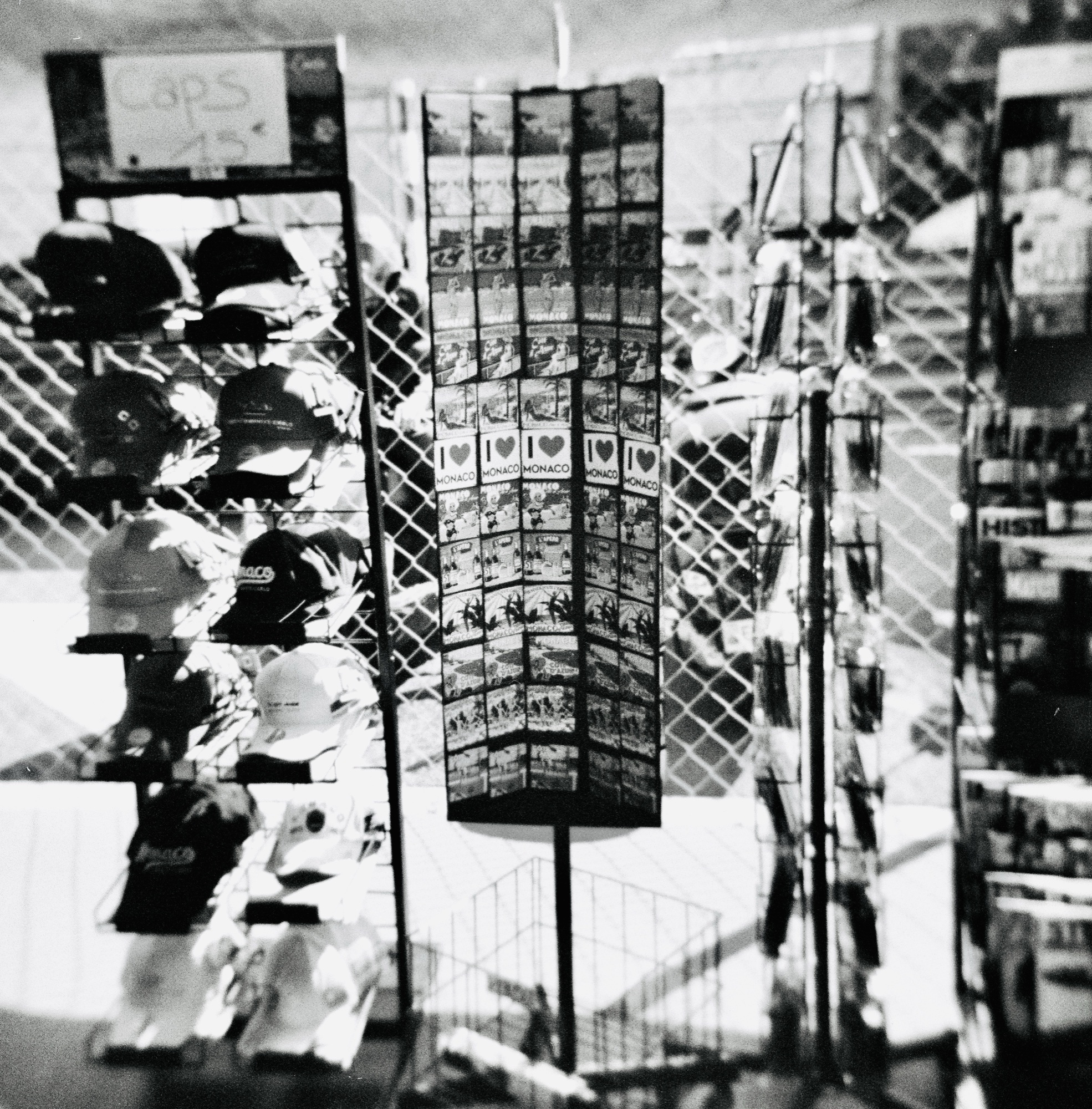 Postcard rack of MONACO souvenirs and caps in front of a chain-link fence, double exposure.